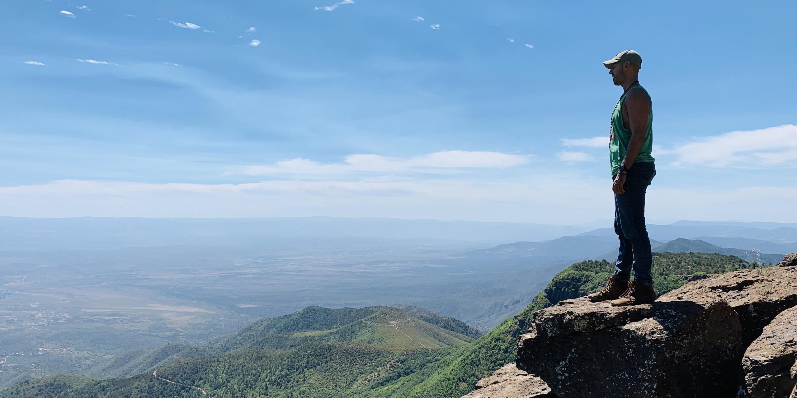 A person standing on a rock ledge high above a wide green valley.