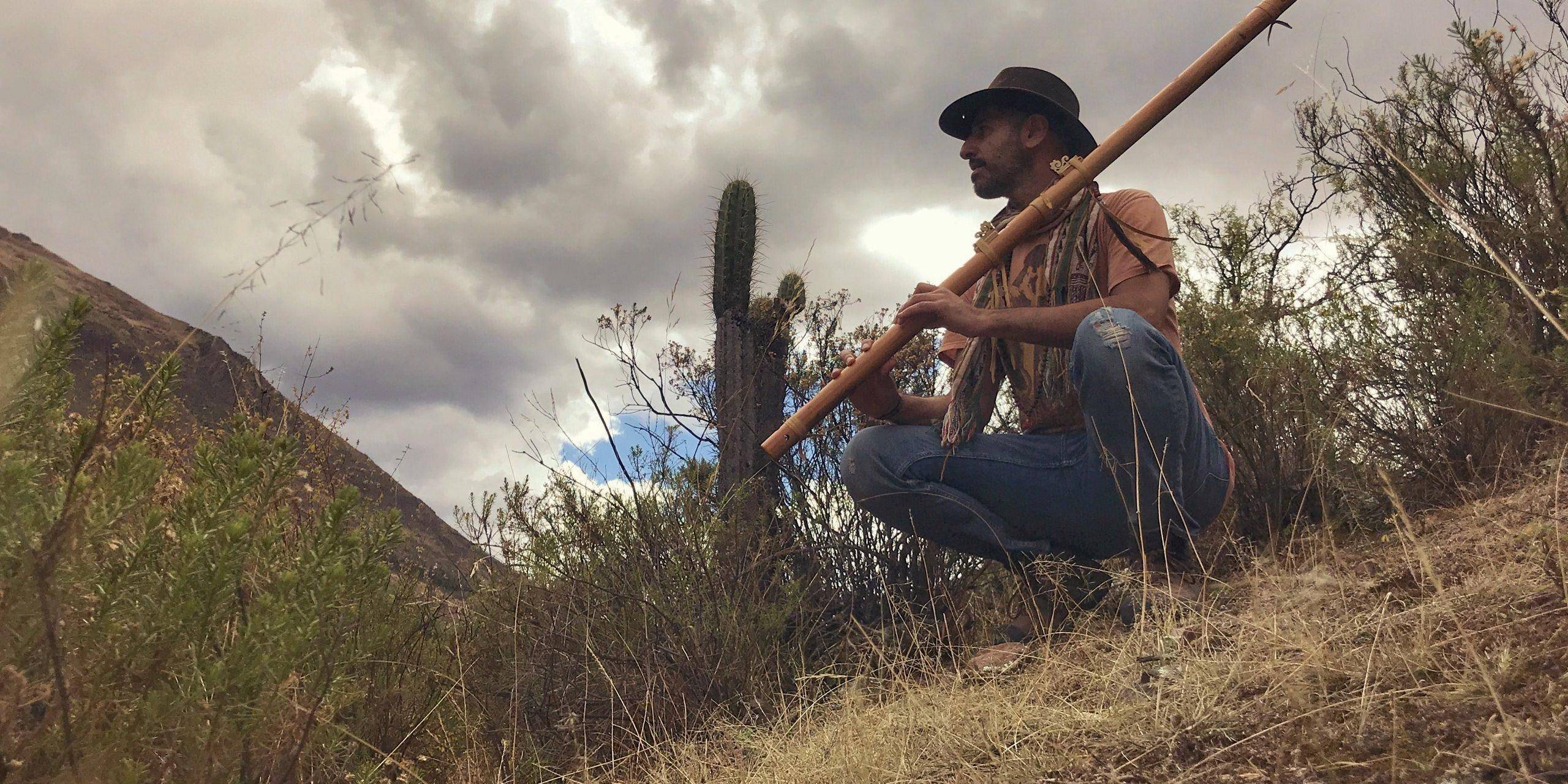 A man in a wide-brimmed hat sits on a hillside playing a long flute.