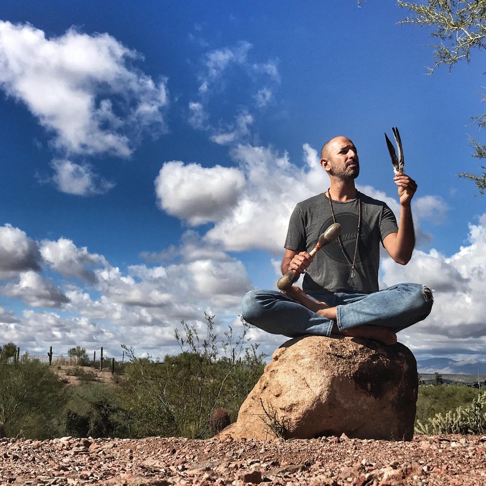 A man seated cross-legged on a boulder holds up a rattle and feathers under a sky of clouds.