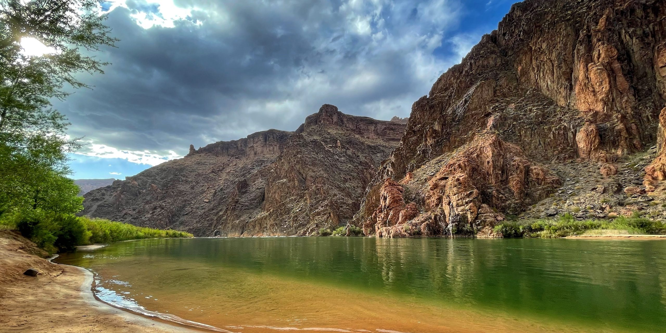 A river running green below red canyon walls, a sandy shore in the foreground.