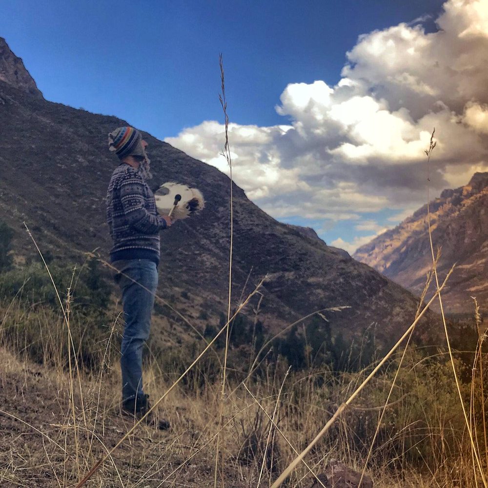 A person holding a frame drum above a wide valley, ridges receding behind them.