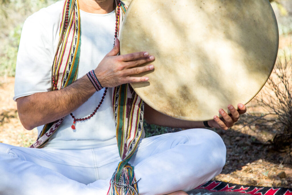 Close view of two hands holding a large frame drum, wearing woven cords and beads, in bright desert light.