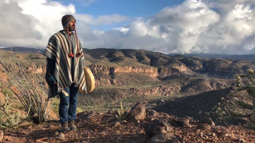 A person in a striped poncho stands at the rim of a wide desert canyon holding a hand drum, with layered cliffs and heaped white cloud behind.