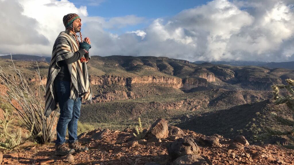 A person in a striped poncho and knitted cap stands with hands together at the rim of a desert canyon under a broad cloudy sky.