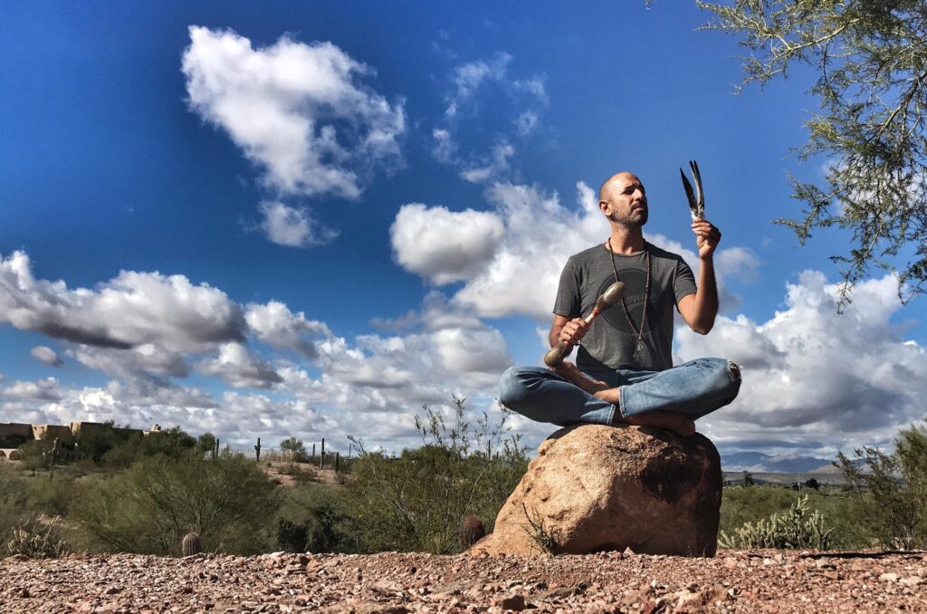 A person sits cross-legged on a boulder holding a rattle and a small drum, under tall white cloud in a blue sky.