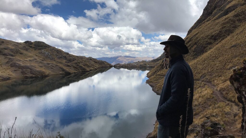 A person in a wide-brimmed hat stands above still green water that mirrors the cloud, held between two steep dry hillsides.