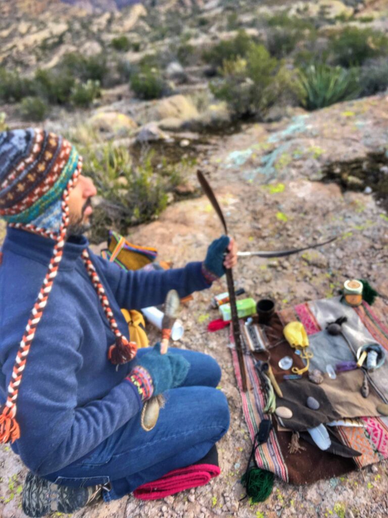 A person in a knitted cap sits cross-legged on a cloth spread with ceremonial objects, on open rock in the desert.
