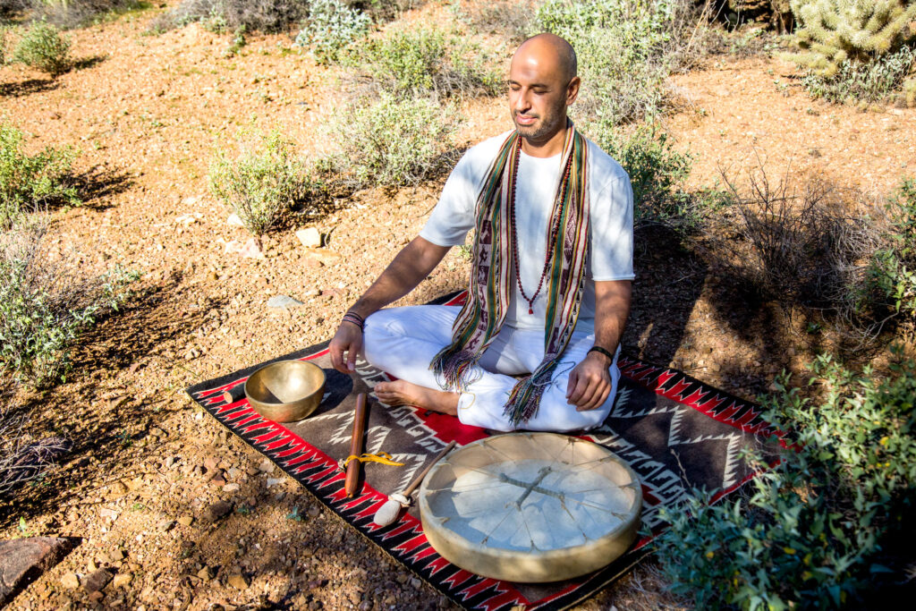 A man in white sits cross-legged on a patterned rug laid on the desert ground, with singing bowls before him.