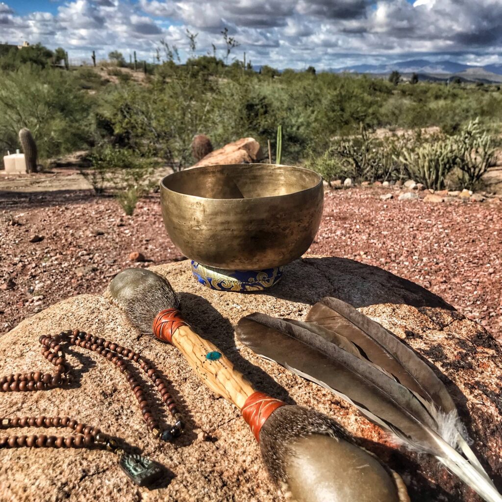 A brass singing bowl, a gourd rattle, a strand of prayer beads and two feather fans laid out on desert ground, with saguaro behind.