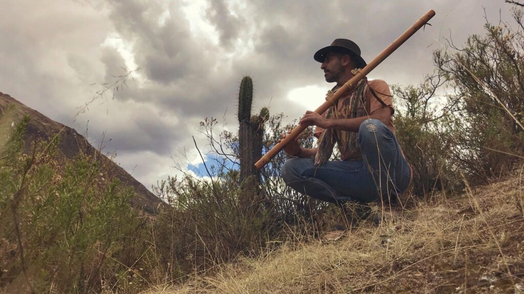 A man in a wide-brimmed hat sits on a hillside playing a long flute.